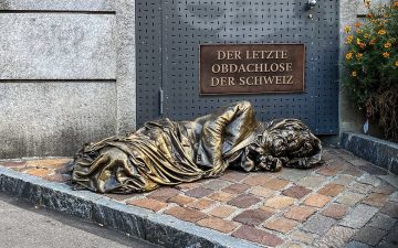 Statue eines Obdachlosen. Oberhalb der Statue steht an der Wand der Schriftzug 